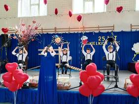 Pokaż zdjęcie: Na scenie trwa występ artystyczny dzieci. Siedzą na krzesłach i i trzymają w dłoniach kapelusze uniesione w górę. 