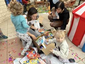 Pokaż zdjęcie: Dziewczynka w blond włosach uczesana w dwa warkoczyki uśmiecha się trzymając pudełko kredek. Kuca obok dużego pudła w którym znajdują się kolorowe przybory plastyczne. Obok stoi druga dziewczynka. Na podłodze leży kolorowe konfetti.