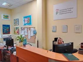 Pokaż zdjęcie: Przy biurku siedzi młoda kobieta, która rozmawia przez telefon. Na ścianie wisi tablica z napisem: Państwowy Fundusz Rehabilitacji Osób Niepełnosprawnych. Ponadto wiszą różne, kolorowe obrazki. 