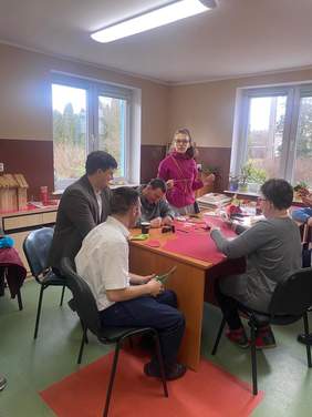 Pokaż zdjęcie: Kilka osób przy stole wycina serca, po lewej stronie zdjęcia stoi mężczyzna