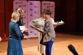 Pokaż zdjęcie: Na scenie Sali Koncertowej stoją cztery osoby: trzy kobiety i mężczyzna. Jedna pani otrzymuje z rąk pozostałych osób prezenty i dyplom. Wszyscy się uśmiechają. 