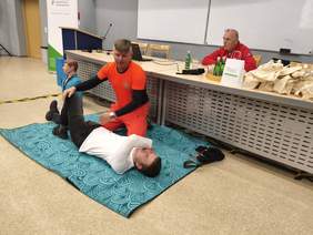 Mężczyzna w pomarańczowym stroju ratownika medycznego klęczy obok młodego chłopaka, trzyma go za zgięte kolano