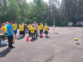 Pokaż zdjęcie: Grupa osób w żółtych koszulkach bierze udział w zawodach sportowych na świeżym powietrzu. Jedna z uczestniczek kopie piłkę.