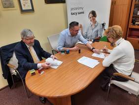 Pokaż zdjęcie: W gabinecie stoi słół, przy którym siedzą trzy osoby: kobieta i dwóch mężczyzn. Jeden z panów pokazuje coś palcem na dokumencie. Na ścianach wiszą różne dyplomy. 