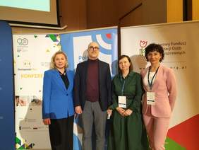 Pokaż zdjęcie: Do pamiątkowego zdjęcia pozują trzy kobiety oraz mężczyzna w okularach. Osoby te stoją na tle baneru z logo PFRON.