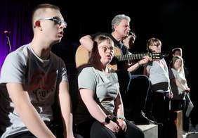 Pokaż zdjęcie: Na ciemnej scenie, ubrani na czarno i szaro stoi pięć osób, w tym jeden mężczyzna grający na gitarze. Osoby te śpiewają.
