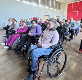 Pokaż zdjęcie: Zdjęcie grupy osób siedzących w sali na krzesłach i wózkach. Na pierwszym planie znajduje się kobieta na wózku, w jasnoróżowym swetrze z ciemnym golfem i w niebieskich spodniach. Włosy ma związane opaską i trzyma ręce splecione na kolanach, patrząc w stronę sceny. Za nią i obok siedzi kilkanaście osób, część również na wózkach - większość patrzy w stronę sceny. Sala ma drewnianą podłogę, jasne ściany, rząd wysokich okien po lewej stronie oraz duży czerwony panel na prawej ścianie. 