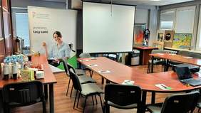 Sala konferencyjna przygotowana na spotkanie informacyjne - stoły ustawione w literę "U", na nich znajdują się notesy i długopisy oraz laptop, z prawej strony ustawiono dwa stoły, na których są napoje, ciasto i naczynia. W tle stoi baner PFRON, obok ekran, a po prawej stronie, oparte na parapecie, prace plastyczne