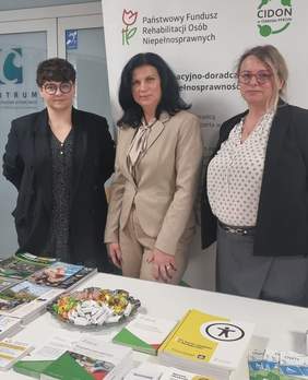 Pokaż zdjęcie: Trzy kobiety stoją przy stoisku informacyjnym. Na stoliku leżą informatory i ulotki. 