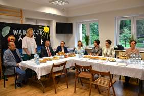 Pokaż zdjęcie: Przy stole siedzą uczestnicy biorący udział w zakończeniu roku szkolnego. Pani Dyrektor Ośrodka wita gości. W tle wakacyjne dekoracje.