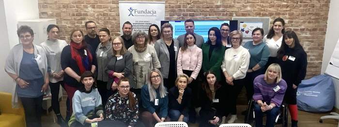 Pokaż zdjęcie: Grupowe zdjęcie kilkudziesięciu osób podczas spotkania Fundacji Aktywizacja. Uczestnicy stoją i siedzą w sali z ceglaną ścianą, banerem fundacji i stołem z notatkami. Część osób ma identyfikatory.