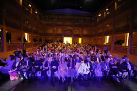 Pokaż zdjęcie: Na dużej wysokiej sali teatralnej w rzędach siedzą goście. Wszyscy elegancko ubrani. Światło jes zaciemnione.