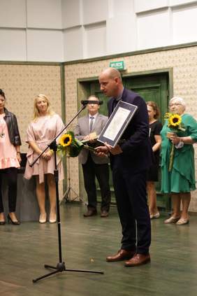 Pokaż zdjęcie: Przed mikrofonem stoi krótko obcięty mężczyzna ubrany w ciemny garnitur. W lewej ręce trzyma dyplom a w prawej kwiaty. Z tyłu za mężczyzną stoją poprzedni laureaci. 