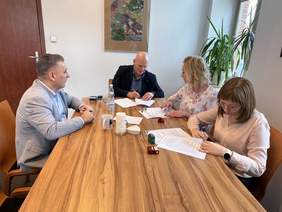 Pokaż zdjęcie: Cztery osoby siedzą przy drewnianym stole w jasnym biurze i podpisują dokumenty. Na stole leżą papiery, pieczątki, kubki, woda i szklanki. W tle widoczne są rośliny i obraz na ścianie.