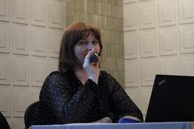 Pokaż zdjęcie: Kobieta z włosami do ramion w rudym kolorze, trzyma blisko ust mkrofon. Siedzi przy stole konferencyjnym i spogląda w laptopa.