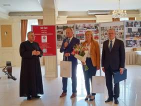 4 osoby, od lewej ksiądz w sutannie obok mężczyzna z mikrofonem następnie kobieta i mężczyzna, stoją w szeregu, jasna sala