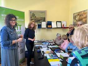 Pokaż zdjęcie: Na stole leżą różne sprzęty, poradniki, informatory. Po prawej stronie siedzą starsi ludzie, którzy przyglądają sie prezentowanym materiałom. Po lewej stronie stoją dwie kobiety, które prowadzą prezentację. 