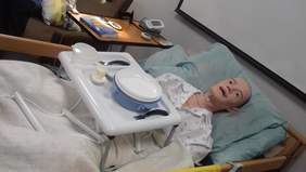 Pokaż zdjęcie: Manekin przypominający osobę starszą, leżący na łóżku rehabilitacyjnym