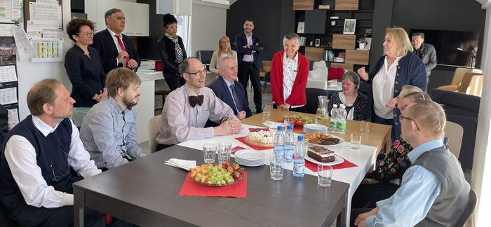 Pokaż zdjęcie: Mężczyzna w garniturze siedzi w przestronnym salonie przy dużym stole, w gronie uśmiechniętych osób. Obok stoi kobieta w blond włosach ciemnej marynarce  która coś mówi. W tle stoją uczestnicy spotkania. Wszyscy ubrani odświętnie.