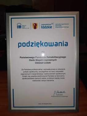 Pokaż zdjęcie: Na stoliku stoją oprawione w ramkę podziękowania dla Łódzkiego Odziału PFRON za profesjonalną i wytrwałą pracę w obszarze polityki społecznej, szczególnie na rzecz obywateli zagrożonych marginalizacją i wykluczeniem społecznym. 