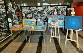 Pokaż zdjęcie: W holu stoją sztalugi, na których ustawione się obrazy i rysunki. Znajduje się także stół, na którym również leżą prace artystyczne. Ilustracje i obrazki są bardzo kolorowe. 