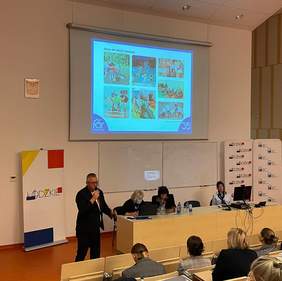 Pokaż zdjęcie: W auli trwa konferencja. Prelegent przemawia do mikrofonu. Na scenie i na widowni siedzą ludzie. 