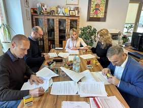 Pokaż zdjęcie: Przy drewnianym dużym stole siedzi pięć osób dwie kobiety w blond włosach i trzej mężczyźni. Na stole leżu mnóstwo dokumentów które panowie podpisują. Za nimi komoda na której stoją kolorowe pamiątki. Po prawej drukarka i komputer na biurk