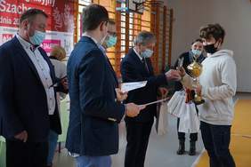 Pokaż zdjęcie: na sali gimnastycznej czterech mężczyzn w garniturach wręcza młodemu chłopakowi puchar statuetkę wraz z dyplomem i upominkiem w tle stoi dziewczyna trzyma białe torby z upominkami