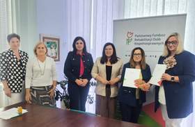 Przy stole konferencyjnym sześć osób pozuje do zdjęcia podczas podpisania umów z PFRON. Dwie osoby trzymają dokumenty, w tle widoczny jest roll-up Funduszu.