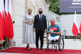 Pokaż zdjęcie: Kobieta i mężczyzna stoją obok sportowca poruszającego się na wózku. W tle budynek pałacu prezydenckiego