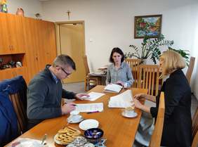 Pokaż zdjęcie: Trzy osoby siedzą przy stole w biurze, przeglądając i podpisując dokumenty. Mężczyzna po lewej stronie, w ciemnym swetrze i okularach, skupia się na podpisywaniu dokumentu. Kobieta po prawej, w czarnym żakiecie, przegląda dokument, a kobieta w środku, w bluzce w kropki, uważnie przygląda się przebiegowi spotkania. W tle widać drewniane szafy, otwarte drzwi, obraz na ścianie oraz duże zielone rośliny.