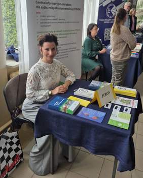 Pokaż zdjęcie: Przy stoliku na którym leżą informatory, foldery, ulotki siedzi młoda kobieta. W tle baner z logo CIDON.