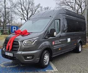 Ciemnoszary bus (duży van) zaparkowany na zewnątrz, z dużą czerwoną kokardą i wstążkami ułożonymi na masce z przodu. Na zdjęciu widać ujęcie z przodu i z boku, z przyciemnianymi oknami w części pasażerskiej. Bus stoi na niebieskim miejscu parkingowym; obok jest znak parkingu z literą „P”, a w tle widać bezlistne drzewa. Na boku pojazdu są logotypy: Miasto i Gmina Grabów nad Prosną oraz Państwowego Funduszu Rehabilitacji Osób Niepełnosprawnych. 
