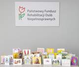 Na jasnoszarym tle u góry znajduje się logo z czerwonym tulipanem i zieloną łodygą oraz napis: Państwowy Fundusz Rehabilitacji Osób Niepełnosprawnych Na dole grafiki stoi rząd rozłożonych, kolorowych kartek wielkanocnych. 