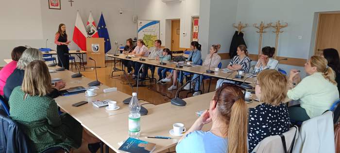 Wokół stołów konferencyjnych siedzą uczestnicy spotkania i słuchają prelegentki, która stoi na przedzie sali. 