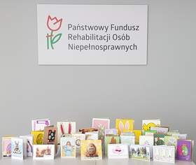 Na jasnoszarym tle u góry znajduje się logo z czerwonym tulipanem i zieloną łodygą oraz napis: Państwowy Fundusz Rehabilitacji Osób Niepełnosprawnych Na dole grafiki stoi rząd rozłożonych, kolorowych kartek wielkanocnych. 