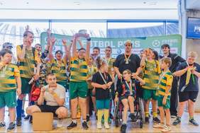 Pokaż zdjęcie: Grupa uczestników turnieju Futbol Plus pozuje do wspólnego zdjęcia przed ścianką wydarzenia. Dzieci i młodzież w strojach sportowych oraz z medalami uśmiechają się i unoszą ręce. Wśród uczestników znajduje się osoba na wózku. Atmosfera radości i świętowania.