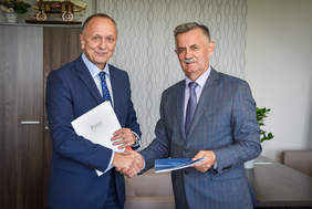 Pokaż zdjęcie: Dwaj mężczyżni, stoją obok siebie, ściskają dłonie, pozują do zdjęcia