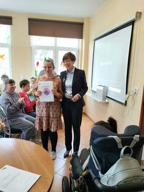 Pokaż zdjęcie: Uczestniczka Warsztatu Terapii Zajęciowej w Wolsztynie prezentuje otrzymany dyplom za udział w konkursie „Sztuka Osób Niepełnosprawnych”. Za nią stoi Anna Skupień, dyrektor Oddziału Wielkopolskiego PFRON. W tle widać grupę osób stojących i siedzących w sali zajęciowej.