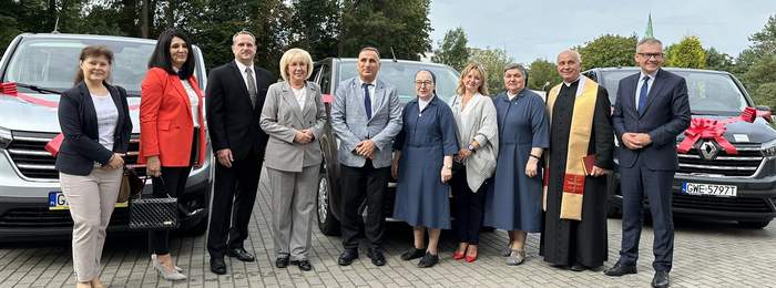 Pokaż zdjęcie: W rzędzie stoi kilka galowo ubranych osób. Wszyscy się uśmiechają. W tle samochody.