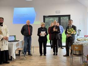 Pokaż zdjęcie: Grupa osób stoi w jasnej sali podczas uroczystości. Po lewej duchowny przy stole, na ścianie plansza z napisem „Fundacja Dom Rain Mana”. W centrum kobieta czyta dokument, obok uczestnicy wydarzenia, drzwi prowadzą na zielony teren.