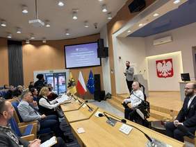 Pokaż zdjęcie: Spotkanie w sali konferencyjnej, gdzie omawiana jest ustawa o asystencji osobistej osób z niepełnosprawnościami. Na pierwszym planie widoczny jest uczestnik na wózku, mówca przy mównicy i symbole Polski, UE oraz regionu.