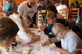 Pokaż zdjęcie: W Sali przy stole siedzi grupa osób. Wszyscy wykonują prace plastyczne. Osoba z zespołem downa przykleja patyczki na kartkę. Kobieta w białej bluzie i białych okularach Szkicuje na kartce papieru. stół jest pokryty białą ceratą w kropki. na sto