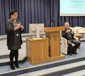 Pokaż zdjęcie: Na zdjęciu widać scenę podczas konferencji dotyczącej Centrów Informacyjno-Doradczych dla Osób z Niepełnosprawnością (CIDON) oraz programów wsparcia PFRON.  Kobieta w czarnej stylizacji z czerwonymi okularami trzyma mikrofon i przemawia do publiczności. Ma na sobie skórzaną kurtkę z frędzlami oraz długi naszyjnik. Obok niej, na podium z godłem Polski, stoi komputer i szklanka wody.  Po prawej stronie siedzi mężczyzna w czarnym stroju, który wykonuje gesty, prawdopodobnie tłumacząc wypowiedź na język migowy. W tle znajduje się ekran z prezentacją oraz granatowa zasłona. Sala wygląda na profesjonalnie przygotowaną do wydarzeń o charakterze edukacyjnym lub informacyjnym.