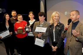 Pokaż zdjęcie: Grupa siedmiu osób stoi na scenie. Jeden z mężczyzn trzyma w ręce mikrofon, a pozostałe osoby trzymają w rękach nagrody rzeczowe, statuetki oraz dyplomy.