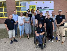 Pokaż zdjęcie: Grupa czternastu osób, wśród których jest także jeden mężczyzna poruszający się na wózku, znajduje się przed budynkiem wykonanym z cegły. W tle widnieje baner Gminy Lisków z herbem gminy.