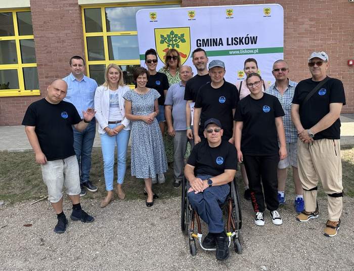 Pokaż zdjęcie: Grupa czternastu osób, wśród których jest także jeden mężczyzna poruszający się na wózku, znajduje się przed budynkiem wykonanym z cegły. W tle widnieje baner Gminy Lisków z herbem gminy.