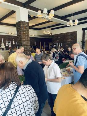 Pokaż zdjęcie: Wokół wielkanocnych stołów zgromadzeni są ludzie. Częstują się światecznymi potrawami. Stoły są pięknie udekorowane wielkanocnymi ozdobami. 