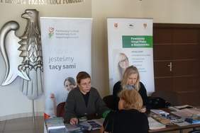 Pokaż zdjęcie: Przy stole na którym są rozłożone broszury i ulotki siedzą dwie kobiet za nimi ustawione są dwa banery PFRON i PUP. Kobieta w okularach udziela informacji kobiecie siedzącej na przeciwko.