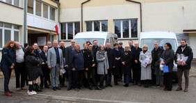 Pokaż zdjęcie: Zdjęcie grupowe wykonane na zewnątrz przed budynkiem. Na zdjęciu widać kilkanaście osób stojących w jednym rzędzie, ubrane w płaszcze i kurtki. W tle stoją dwa srebrne busy dla Warsztatów Terapii Zajęciowej w Grodzisku Wielkopolskim i Rakoniewicach.